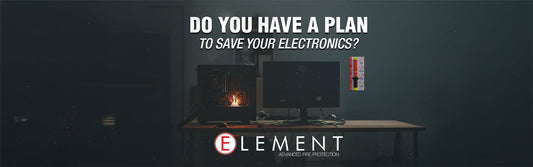 An office setup where a small fire has ignited inside a desktop computer tower sitting on a wooden desk. Next to the tower is a computer monitor with various cables visible. An Element Fire extinguisher is mounted on the wall behind the desk.